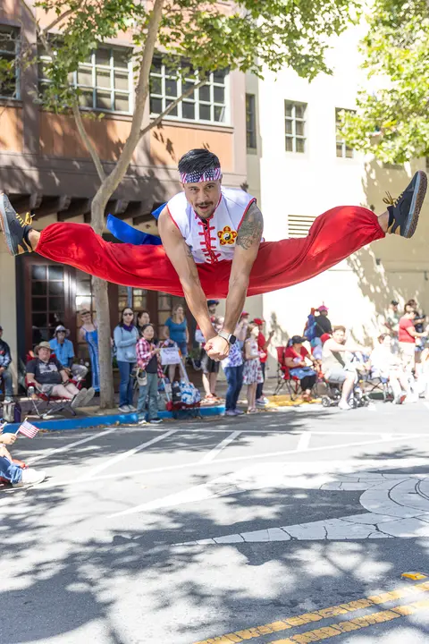 A performer does a high jump split in the middle of a street during an outdoor event.