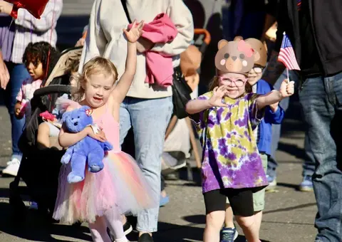 Children participate in a parade, waving and holding toys.