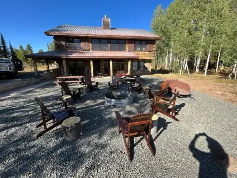 A rustic log cabin with an outdoor fire pit and wooden chairs in a gravel yard.