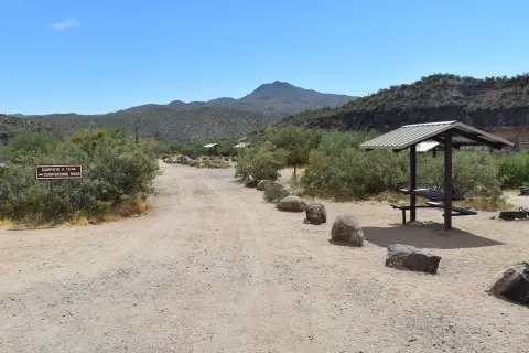 A dirt road leads through a desert campground with picnic shelters and mountains in the background.