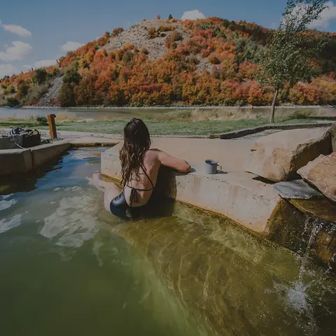 A woman relaxes in an outdoor hot spring overlooking a scenic landscape with autumn trees.