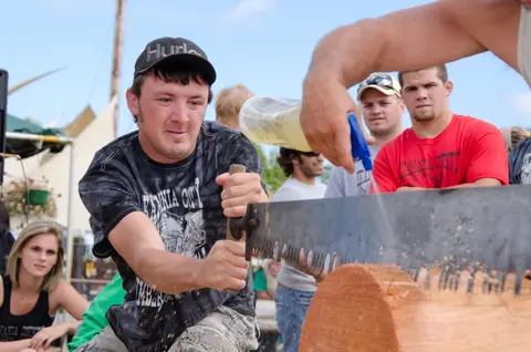 A man uses a crosscut saw to cut a log while another person sprays a lubricant.