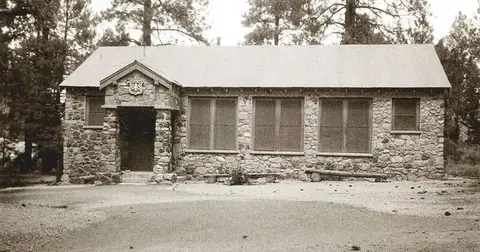 A historic black and white photo shows the exterior of a stone building surrounded by trees.