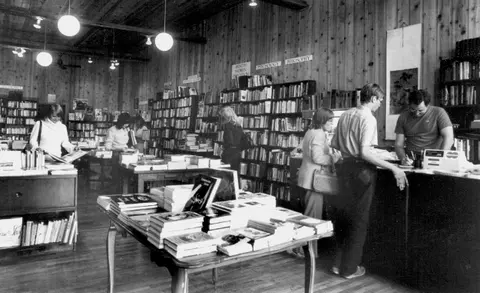 Black and white photo of the interior of a bookstore with people browsing and checking out.
