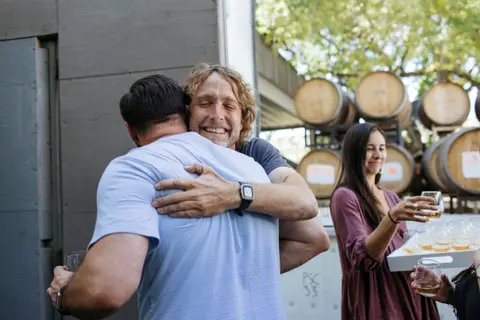 Two men embrace outdoors near wine barrels while a woman stands nearby holding a glass.