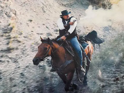 A man rides a horse while firing a revolver.