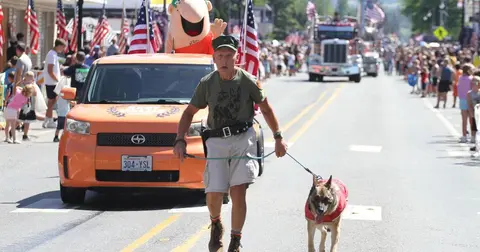A man walks his dog in front of an orange car in a parade.