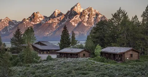 A scenic view of wooden cabins in front of snow-capped mountains and evergreen trees.