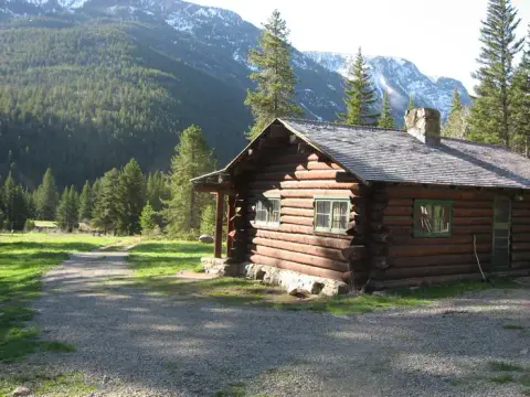 A log cabin sits near a dirt path with mountains in the background.