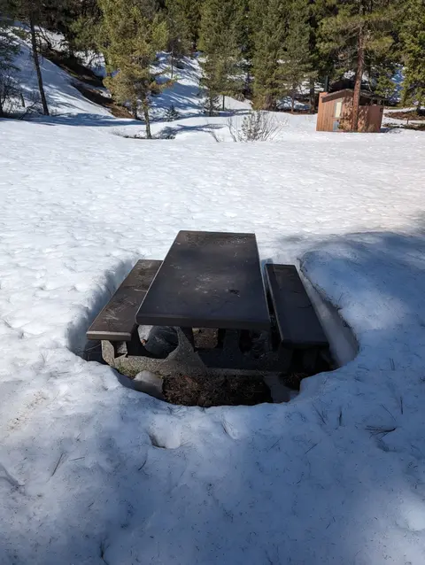 A picnic table sits in a snowy landscape with a small building in the background.