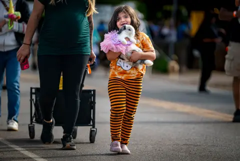 A young girl happily carries a bunny wearing a pink tutu while walking down a street.