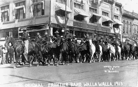 A historic black and white photograph showing a band on horseback riding down a city street.