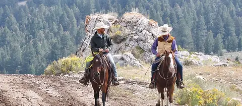 Two cowboys ride horses along a dirt trail with rocky outcrops and a forest in the background.