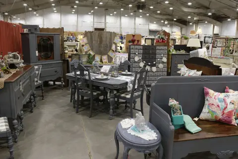An indoor exhibition hall displaying various painted wooden furniture sets and home decor items.