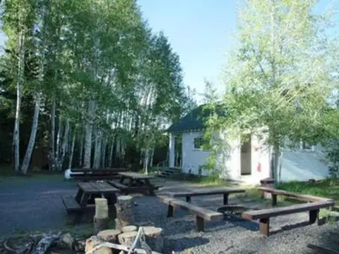 A building stands surrounded by tall trees with outdoor picnic tables and a fire pit.