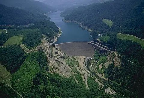 An aerial view of a large dam and reservoir surrounded by forested mountains.