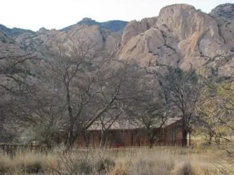A rustic building nestled among bare trees below rugged mountains.