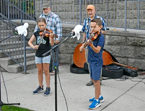 A young girl and boy play violins outdoors while adults watch in the background.