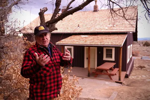 A man wearing a red plaid jacket stands outside a wooden cabin with a shingle roof.