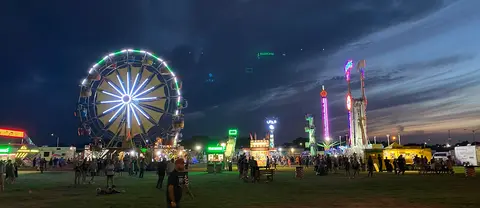 A brightly lit Ferris wheel and carnival rides operating at dusk with people walking about.