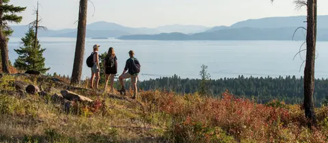 Three people stand on a hillside overlooking a large lake and mountains.