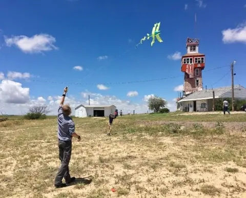 A person flies a green kite in an open field near a tall structure.