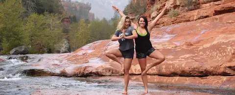 Two women doing yoga poses on red rocks by a stream.