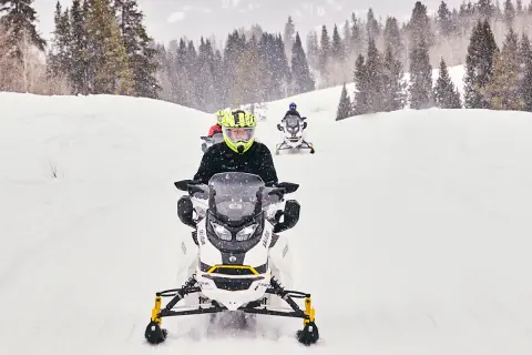 People ride snowmobiles on a snowy trail in the woods.