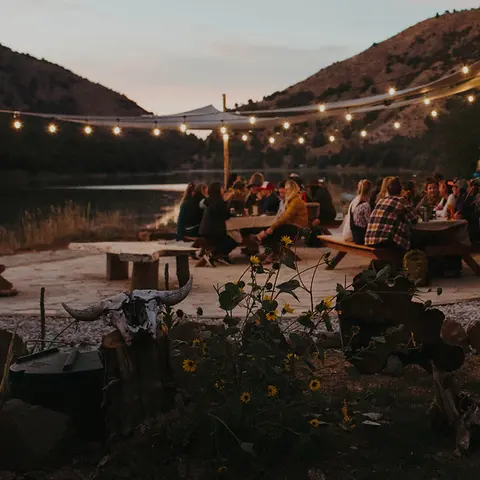 People gathered outside for a meal at dusk near a body of water and hills.