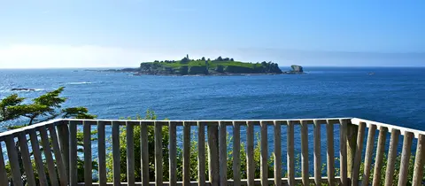A view of a grassy island with a lighthouse from behind a wooden railing over the ocean.
