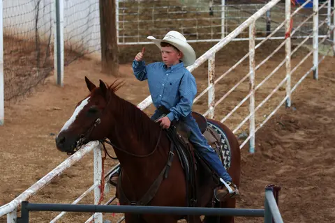 A young boy in a cowboy hat sits on a horse in an arena, giving a thumbs up.