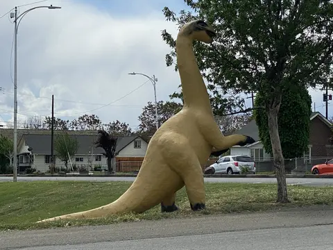 A large dinosaur statue stands in a grassy area near a street and houses.