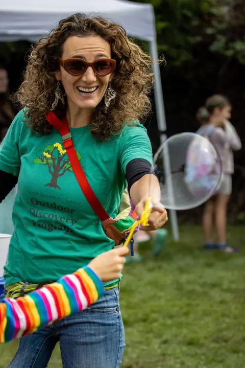 A smiling woman wearing sunglasses holds a bubble wand outdoors.