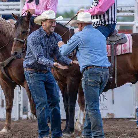 Two cowboys interact while standing near horses at an outdoor arena.