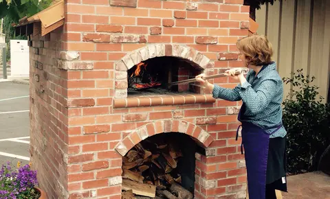 A woman tends a fire inside a brick pizza oven outdoors.
