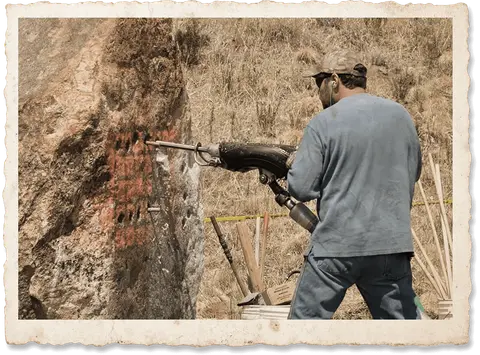 A man uses a large drill on a large rock outdoors.
