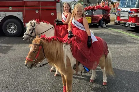 Two children in red dresses ride ponies in front of fire trucks.