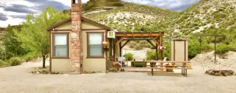 A small cabin with a stone chimney sits in a desert landscape with hills behind it.