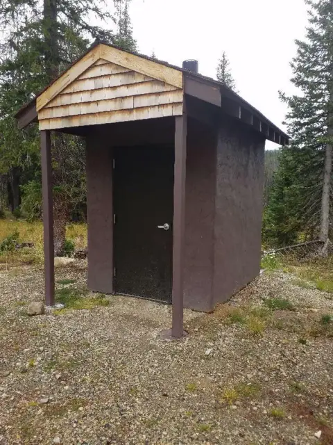 A small brown outhouse stands in a clearing surrounded by trees.