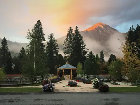 A wooden gazebo sits amid flower planters and trees with a mountain and sunrise sky in the background.