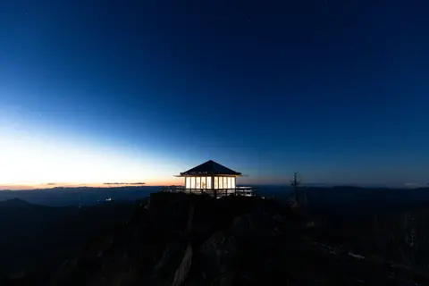 A brightly lit lookout cabin sits atop a dark rocky mountain peak at twilight.