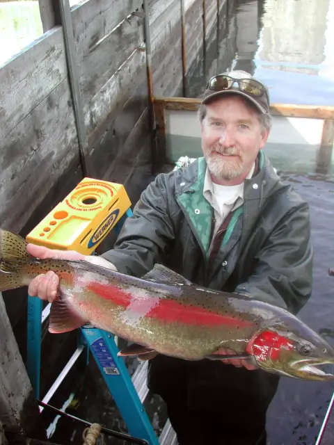 A smiling man holds a large fish he caught near the water.
