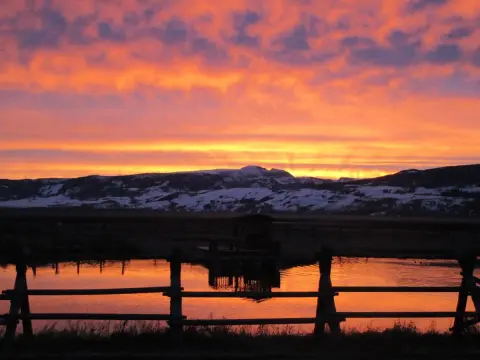 A vibrant sunset over a snowy mountain range with a wooden fence and pond in the foreground.