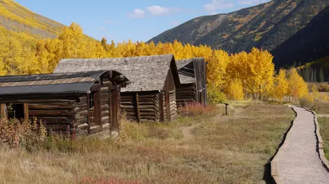 Wooden cabins stand alongside a path with mountains and yellow trees in the background.