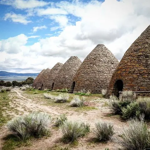 A row of stone beehive kilns stand in an open landscape under a partly cloudy sky.