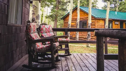 Two rustic rocking chairs on a wooden porch with a cabin and aspen trees in the background.