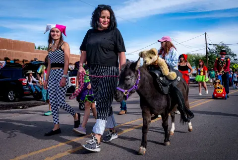 People in costume walk down a street leading a pony with a stuffed bear on its back.