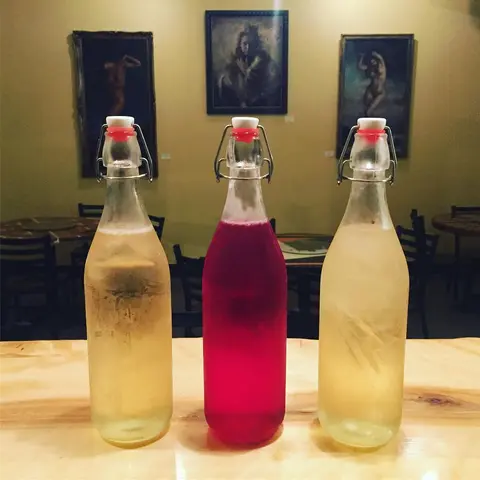 Three swing-top glass bottles filled with beverages stand on a wooden counter in front of framed artwork.