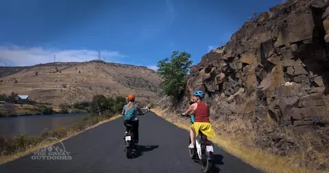 Two people ride bicycles along a paved path next to a rocky cliff and a river.