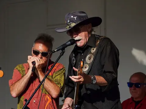 Two musicians perform on stage, with one singing into a microphone and playing guitar while the other plays the harmonica.
